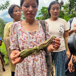 May include: A woman in a floral dress holds a bundle of green, knotted plant material. Other women stand nearby, some wearing light-colored shirts. The scene appears to be outdoors, with trees and a cloudy sky in the background.