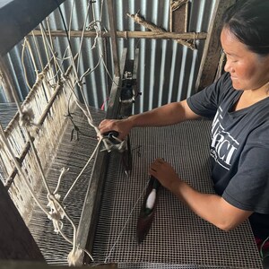 May include: A woman weaving on a wooden loom. The loom is made of wood and has white and gray threads. The woman is wearing a black t-shirt with white text. The woven fabric appears to be a black and white checkered pattern.