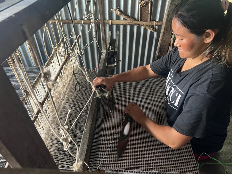 May include: A woman weaving on a wooden loom. The loom is made of wood and has white and gray threads. The woman is wearing a black t-shirt with white text. The woven fabric appears to be a black and white checkered pattern.