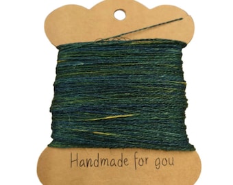 May include: A spool of green and yellow thread, labeled "Handmade for you", is wound around a brown cardboard bobbin. The thread is a mix of dark green and yellow, creating a textured appearance. The bobbin has a scalloped edge and a hole at the top.