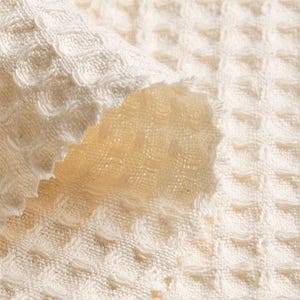 May include: Close-up of a white waffle weave fabric with a textured surface. The fabric is folded over, revealing the underside of the weave.