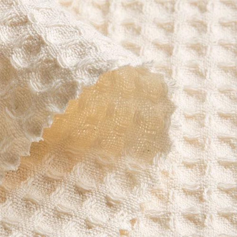 May include: Close-up of a white waffle weave fabric with a textured surface. The fabric is folded over, revealing the underside of the weave.
