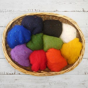 Può includere: Un cesto di vimini pieno di lana cardata colorata nelle tonalità di blu, nero, bianco, verde, viola, rosso, arancione e giallo. La lana ha macchie di colore. Il cesto è appoggiato su una superficie di legno bianco.