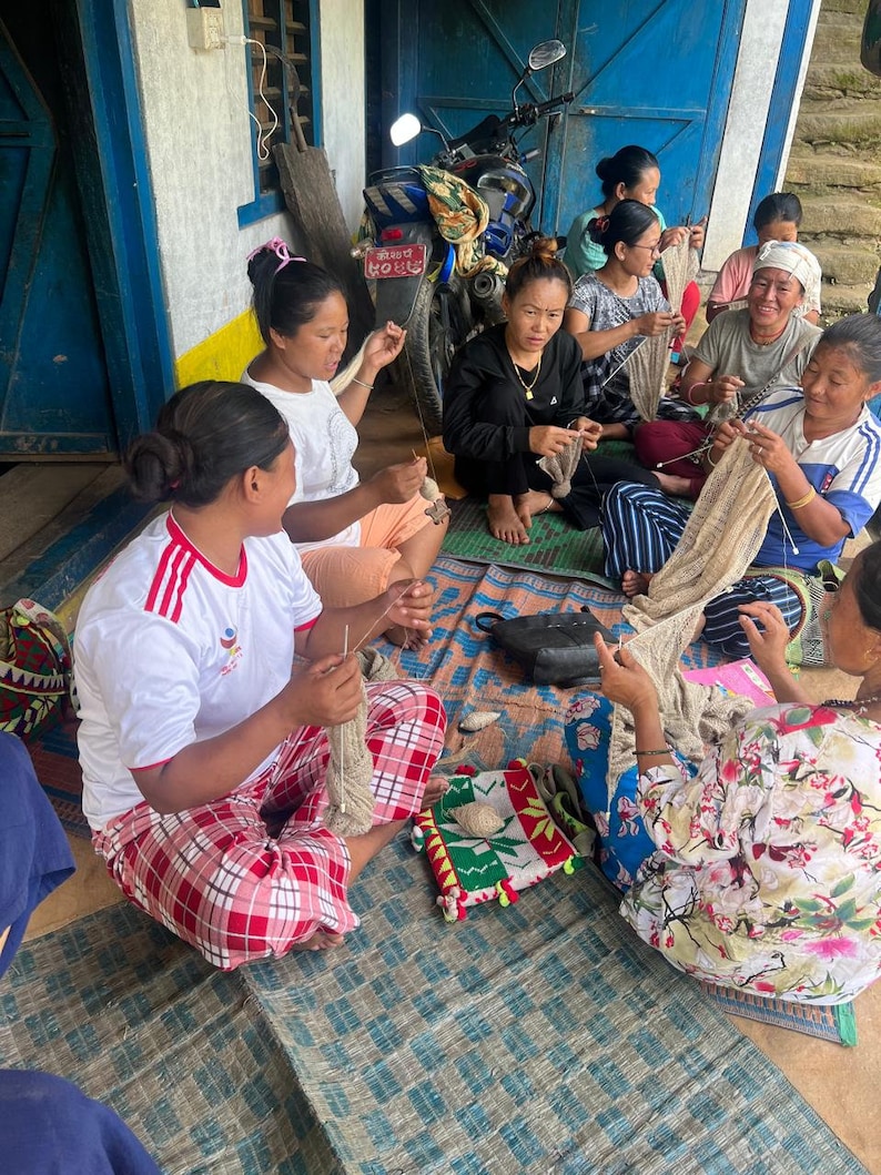 May include: A group of women are gathered, engaged in knitting or weaving. They are seated on a patterned mat, working with yarn and needles. The scene suggests a community craft activity, with various textiles and tools visible.