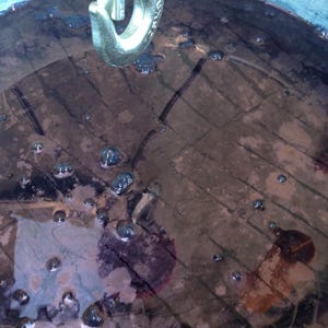 May include: A close-up of a large metal container filled with a dark purple liquid. The liquid is bubbling and there are small air bubbles on the surface. A metal hook is hanging from the top of the container.
