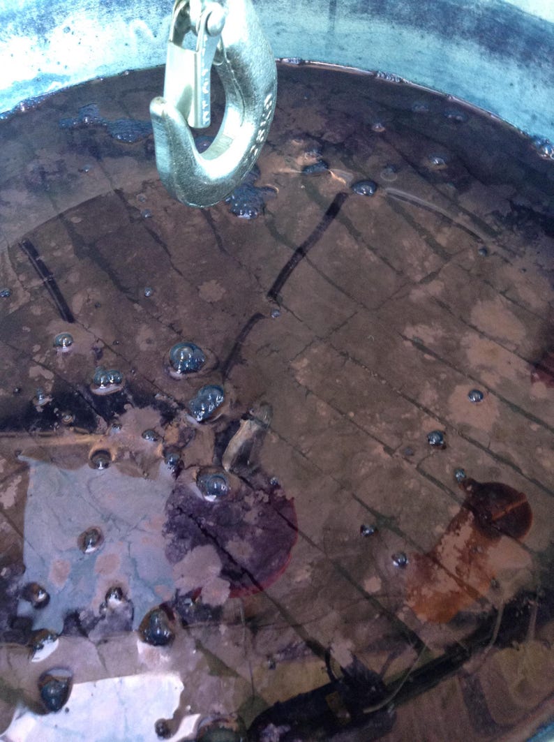 May include: A close-up of a large metal container filled with a dark purple liquid. The liquid is bubbling and there are small air bubbles on the surface. A metal hook is hanging from the top of the container.
