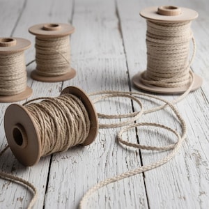 Puede incluir: Varias bobinas de madera con hilo natural dispuestas sobre una superficie de madera blanca desgastada. El hilo es de color beige claro, y parte de él está desenrollado, creando bucles. Las bobinas son de madera marrón claro.