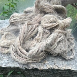 May include: A pile of raw, natural-colored hemp fiber rests on a gray stone surface. The fibers are loosely arranged, showing a textured, organic appearance. The hemp is a light brown color, with some areas appearing lighter.