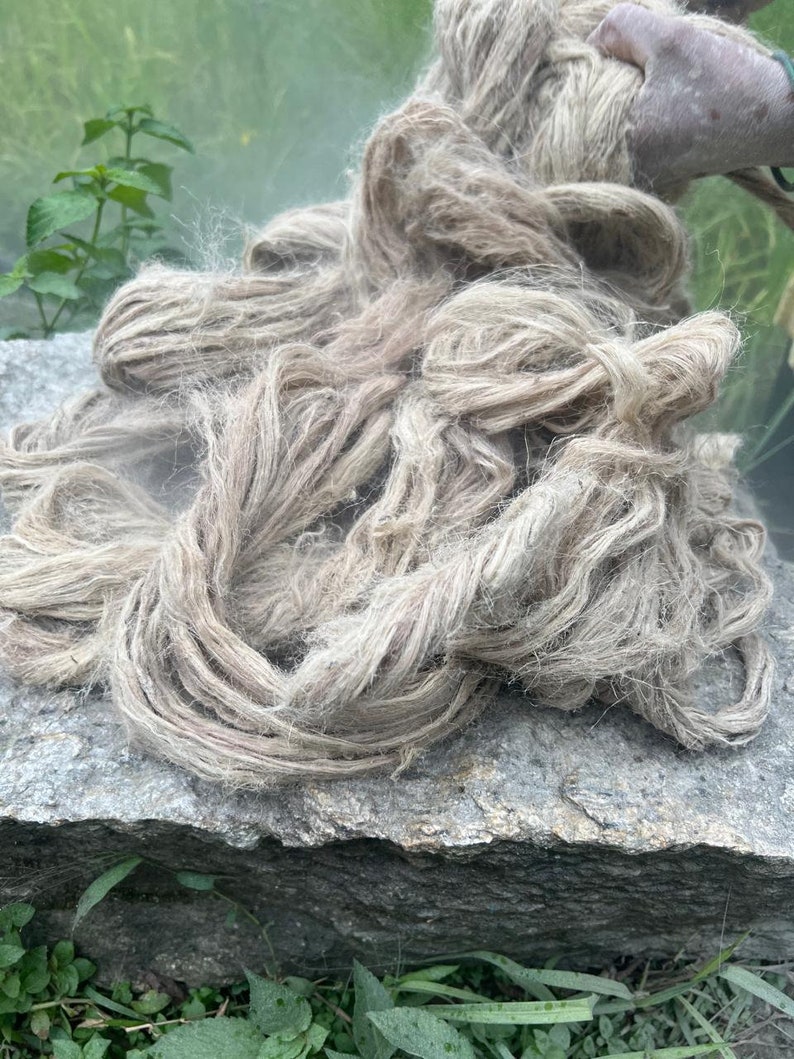 May include: A pile of raw, natural-colored hemp fiber rests on a gray stone surface. The fibers are loosely arranged, showing a textured, organic appearance. The hemp is a light brown color, with some areas appearing lighter.