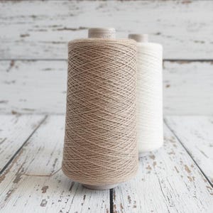 Könnte beinhalten: Zwei Garnspulen, eine in Hellbraun und die andere in Weiß, stehen nebeneinander. Das Garn ist eng um einen konischen Kern gewickelt. Der Hintergrund ist eine verwitterte, weiße Holzoberfläche.