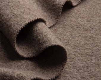 May include: Close-up of a folded section of brown wool fabric. The fabric has a soft, textured appearance with a slightly uneven surface. The edges of the fabric are cut with a scalloped pattern. The overall color is a warm, neutral brown.