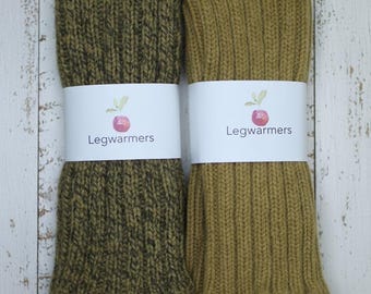 May include: Two pairs of knitted leg warmers in olive green and mustard yellow, each wrapped with a white band featuring the text "Legwarmers" and a small illustration. The leg warmers are displayed on a white wooden surface.
