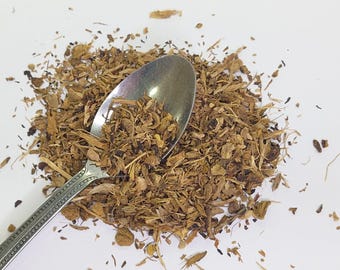 May include: A pile of dried, brown herbs with a silver spoon resting on top. The spoon is angled towards the upper left, and the herbs are scattered across a white surface. The herbs appear to be small pieces of dried roots or bark.