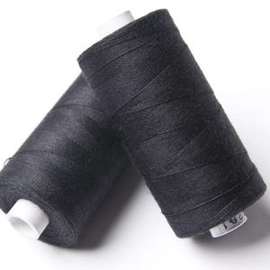 May include: Two spools of black thread are angled towards each other against a white background. The thread is tightly wound around white cardboard cores. The spools are likely for sewing or crafting projects.