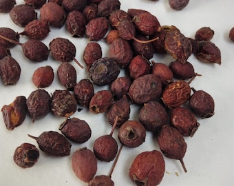 May include: A collection of dried, dark red berries, some with a slightly purple hue, scattered on a white surface.