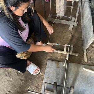 May include: A woman working with a wooden spinning wheel, winding thread. The thread is a light beige color, and the wheel is made of wood. The woman is wearing glasses and a black and pink shirt. The floor is dirt.