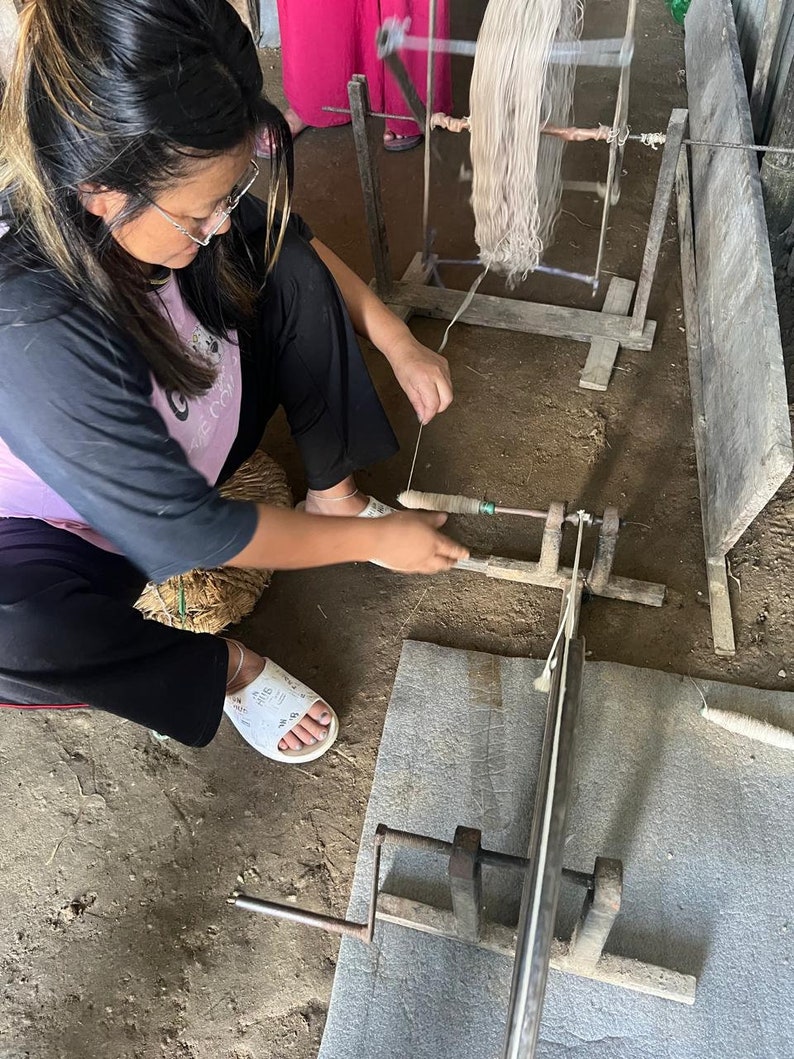 May include: A woman working with a wooden spinning wheel, winding thread. The thread is a light beige color, and the wheel is made of wood. The woman is wearing glasses and a black and pink shirt. The floor is dirt.