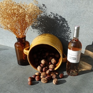 May include: A still life composition with a bottle of rosé wine, a yellow ceramic pot filled with nuts, and a brown glass vase holding dried flowers. The wine bottle has a silver cap and a white label.