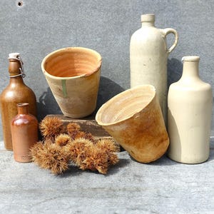 Może przedstawiać: Kolekcja pięciu przedmiotów z ceramiki vintage, w tym dwie brązowe butelki szklane, dwie doniczki z terakoty i dzban z białej ceramiki. Przedmioty są ułożone na szarej, teksturowanej powierzchni.