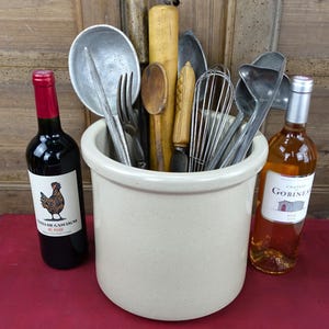 Puede incluir: Un recipiente de cerámica color crema lleno de utensilios de cocina, incluyendo cucharas de madera, batidores y cucharones de metal. Dos botellas de vino, una tinto y otra rosado, se colocan a los lados.