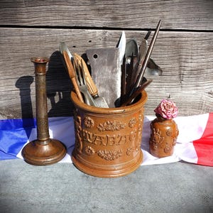 May include: A brown ceramic crock with embossed floral patterns and the word TABAC, filled with various metal kitchen utensils including a cleaver and fork. Beside it stands a tall metal candlestick and a small brown ceramic bottle with a dried flower.