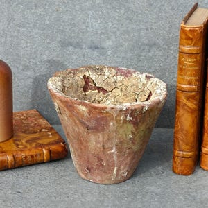 May include: A still life arrangement featuring a brown bottle, a weathered terracotta pot, and two vintage books with leather covers. The objects are set on a grey surface, creating a rustic aesthetic.