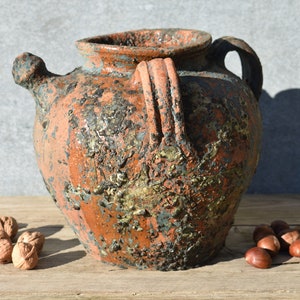 Pu&ograve; includere: Un'anfora in ceramica rustica di colore terracotta con un beccuccio e due manici. L'anfora ha un aspetto screziato e invecchiato con macchie di vernice blu e dorata.