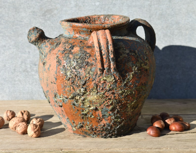 Pu&ograve; includere: Un'anfora in ceramica rustica di colore terracotta con un beccuccio e due manici. L'anfora ha un aspetto screziato e invecchiato con macchie di vernice blu e dorata.