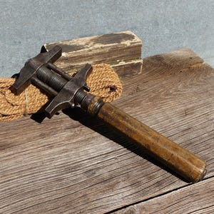 May include: An antique metal clamp with a wooden handle, on a weathered wooden surface. The clamp features a screw mechanism and two flat arms. A coil of rope and a piece of wood are in the background.