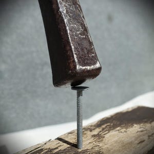 May include: A dark brown, weathered wooden hammer handle is positioned directly above a silver-coloured metal nail. The nail is driven vertically into a piece of rough, light-coloured wood that shows signs of wear and aged texture.