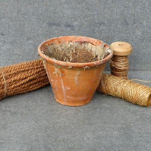 May include: An orange terracotta flower pot with a weathered look, alongside a bundle of brown rope and two spools of twine. The items are arranged on a grey surface, suggesting a gardening or craft theme.