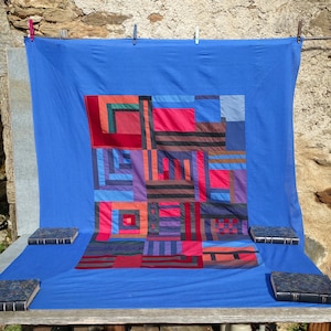 Pode incluir: Uma colcha azul com um padrão geométrico colorido de quadrados e retângulos. A colcha está pendurada em um varal contra uma parede de pedra.