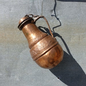 K&ouml;nnte beinhalten: Ein kupferner Krug mit einem verzierten Design h&auml;ngt an einem Haken. Der Krug hat einen runden K&ouml;rper und einen schmalen Hals mit einem Ausguss. Das Design zeigt ein sich wiederholendes Muster aus Dreiecken und Linien.