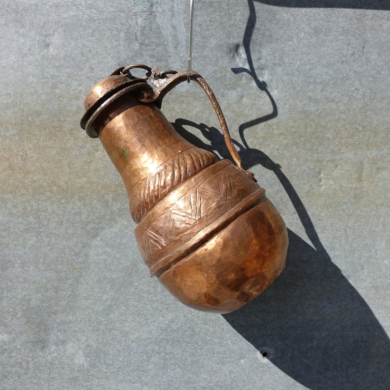 K&ouml;nnte beinhalten: Ein kupferner Krug mit einem verzierten Design h&auml;ngt an einem Haken. Der Krug hat einen runden K&ouml;rper und einen schmalen Hals mit einem Ausguss. Das Design zeigt ein sich wiederholendes Muster aus Dreiecken und Linien.