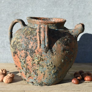 Pu&ograve; includere: Un grande vaso in terracotta rustico con una finitura scheggiata e consumata. Il vaso ha un'ampia apertura e un manico su un lato. Il vaso &egrave; di un colore blu-grigio scuro con macchie arancioni e marroni.
