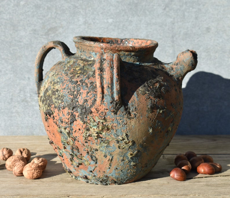 Pu&ograve; includere: Un grande vaso in terracotta rustico con una finitura scheggiata e consumata. Il vaso ha un'ampia apertura e un manico su un lato. Il vaso &egrave; di un colore blu-grigio scuro con macchie arancioni e marroni.