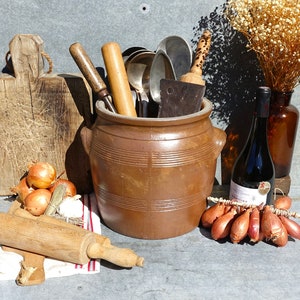 以下が含まれることがあります： 木製の麺棒、茶色の陶器の壺、木製のまな板、赤い玉ねぎの束、赤ワインのボトルなど、ヴィンテージのキッチンツールと食材のコレクションです。