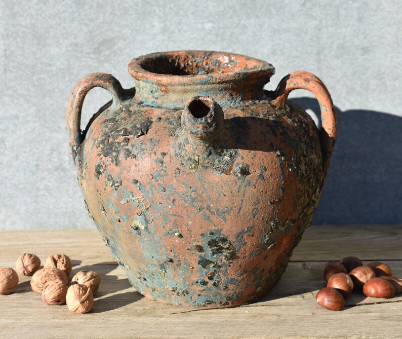 Pu&ograve; includere: Un grande vaso in terracotta rustico con un beccuccio e due manici. Il vaso ha una smaltatura blu e marrone screziata e mostra segni di et&agrave; e usura.