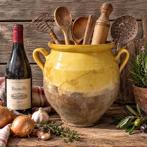Peut inclure: Un grand pot en céramique émaillée jaune et marron avec deux anses, rempli d'ustensiles de cuisine en bois dont un fouet, des cuillères et un rouleau à pâtisserie. À côté se trouvent une bouteille de vin Brouilly, deux oignons, de l'ail et des herbes.