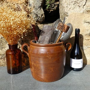 May include: A brown ceramic crock with two handles, filled with kitchen utensils, including a whisk, a ladle, and a wooden spoon. A bottle of wine is next to the crock, and a vase with dried flowers is in the background.