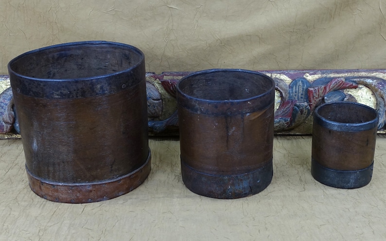 May include: Three brown wooden buckets with metal bands around the top and bottom. The buckets are different sizes and are arranged from largest to smallest.
