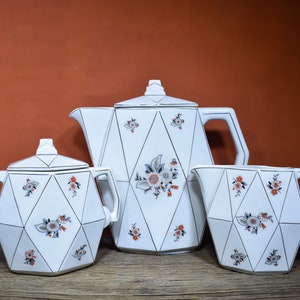 May include: A white ceramic tea set with a geometric design and floral accents. The set includes a teapot, creamer, and sugar bowl.