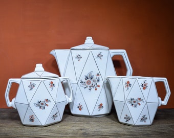 Puede incluir: Un juego de té de cerámica blanca con un diseño geométrico y detalles florales. El juego incluye una tetera, una jarra de crema y un azucarero.