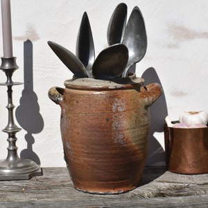 May include: A brown ceramic crock with a wide mouth and two handles, filled with a collection of vintage silver spoons.
