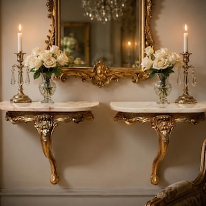 May include: Two matching wall-mounted console tables with gold-toned ornate carved supports and white marble tops. Each table holds a clear glass vase filled with white roses and a tall brass candlestick with a lit white taper candle.