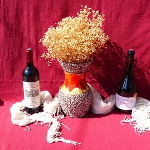 May include: Two bottles of wine, one red and one white, are on a red tablecloth with a white lace doily. A ceramic vase with a brown and orange speckled design holds a bouquet of dried white flowers.