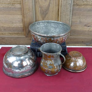 May include: A selection of antique copper kitchenware, including a large bowl, a small jug with a handle, and two smaller bowls. The copper has a weathered appearance, with silver and brown color variations.