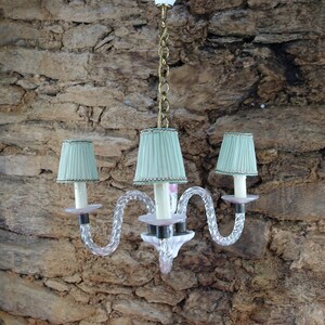 May include: A three-light chandelier with a twisted glass design and light green lampshades. The chandelier is hanging from a chain and has a pink and white glass base.
