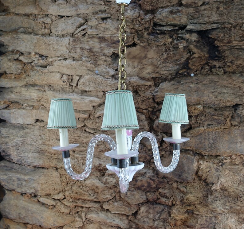 May include: A three-light chandelier with a twisted glass design and light green lampshades. The chandelier is hanging from a chain and has a pink and white glass base.