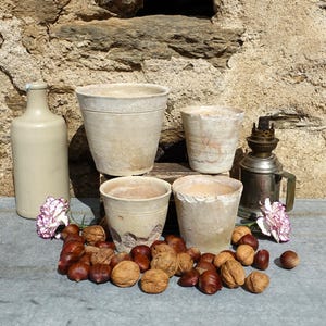 Może przedstawiać: Cztery doniczki z terakoty w różnych rozmiarach, z rustykalnym, postarzanym wykończeniem, są ustawione na stole z butelką i lampą. Doniczki są otoczone kasztanami i różowymi kwiatami.